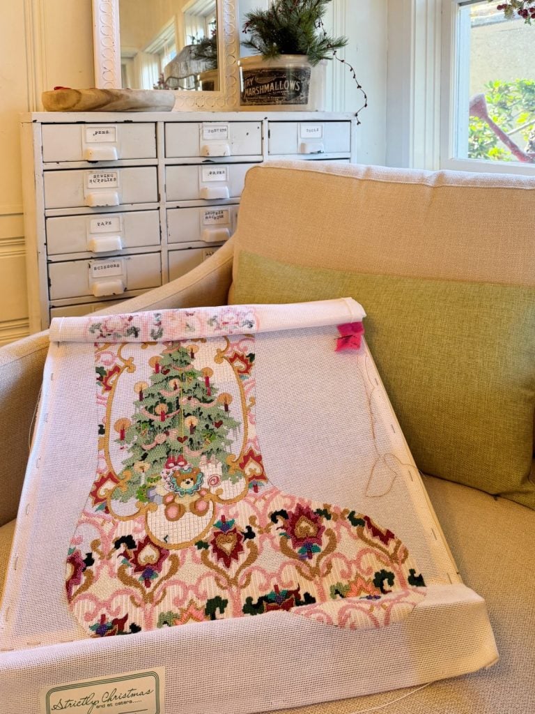 A partially completed cross-stitch showing a detailed Christmas stocking design is placed on a beige armchair, with a cabinet and festive greenery in the background.