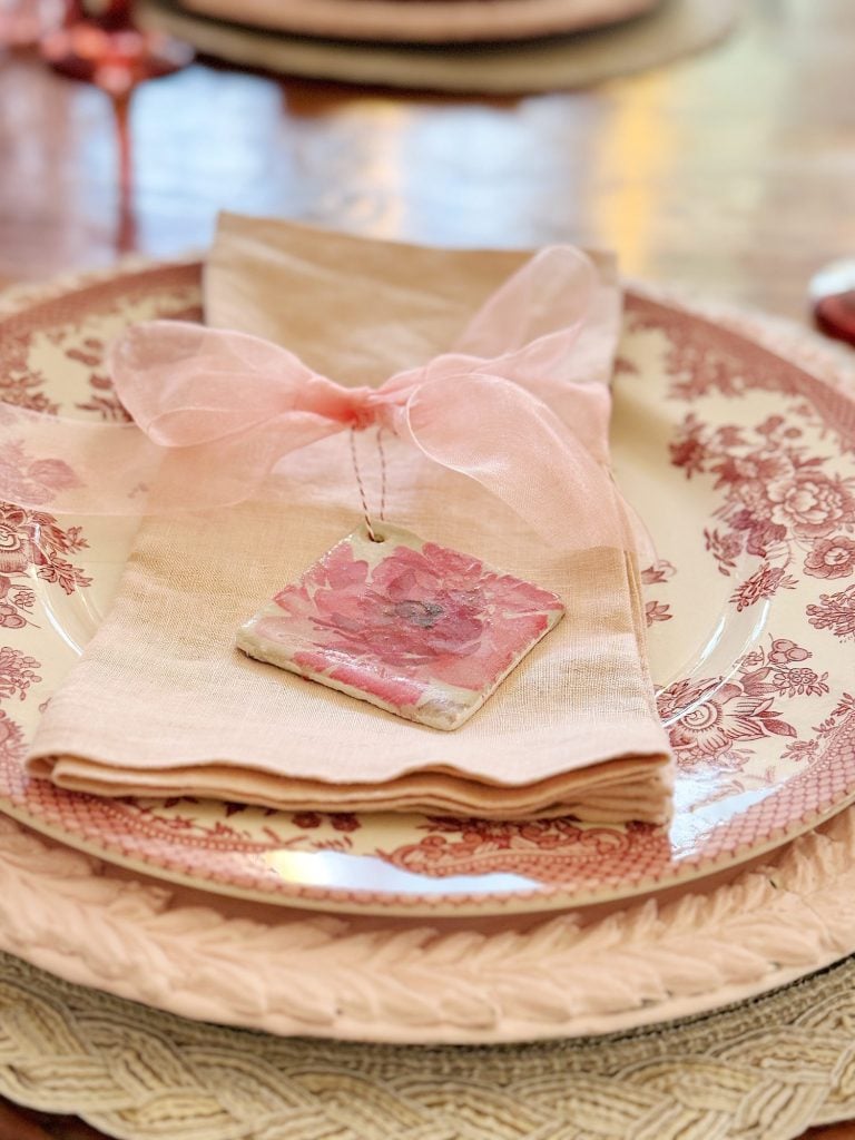 A decorative table setting with a floral-patterned plate, beige napkin tied with a pink ribbon, and a small floral tile.