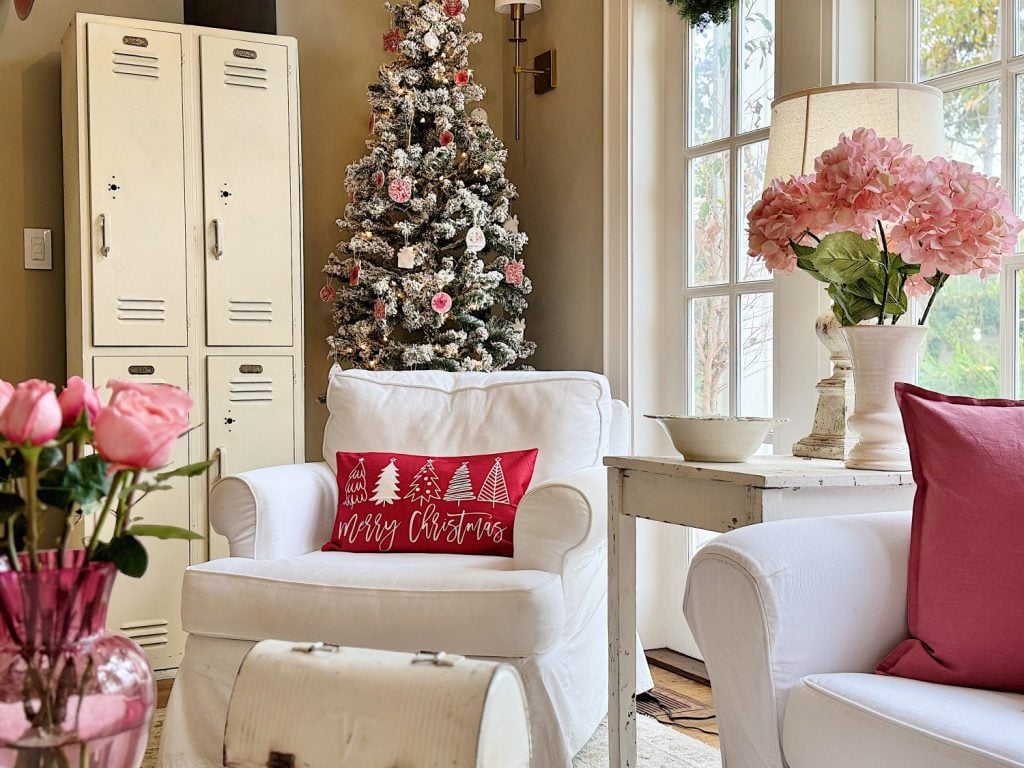 A cozy living room with a white armchair, pink flowers, and a snowy Christmas tree. A red pillow with "Merry Christmas" is on the chair. White lockers are next to the tree.