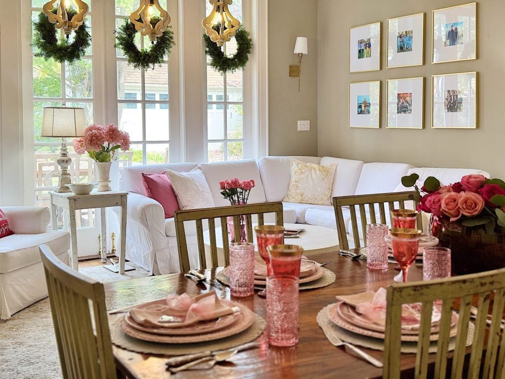 A dining and living room with a wooden table set with pink glasses and plates, white sofas, floral centerpiece, and decor. Framed photos and wreaths adorn the walls and windows.