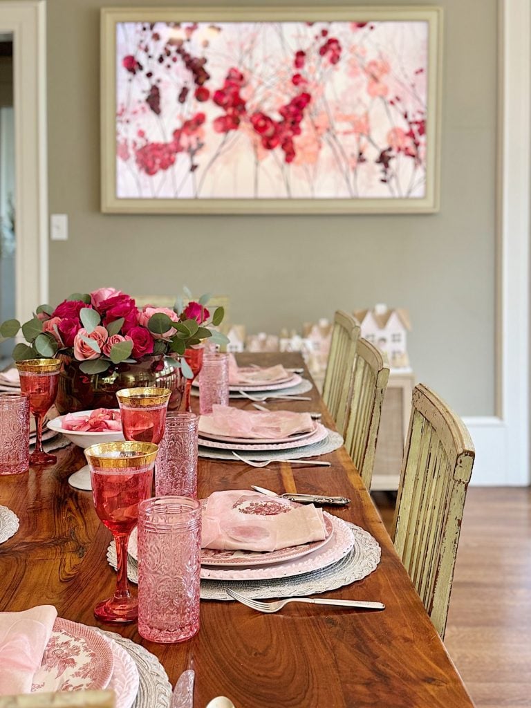 A dining table is set with pink glassware, floral plates, and a centerpiece of red and pink flowers. A large abstract floral artwork hangs on the wall.