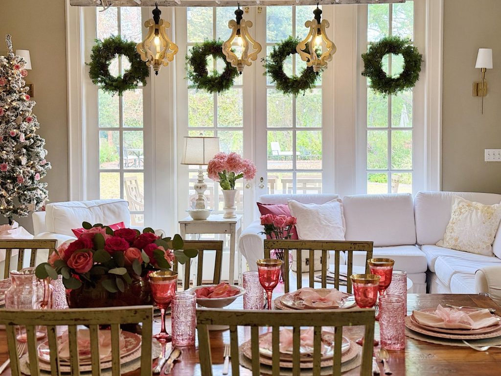 A festive dining room with a table set for a meal. Red and pink flowers are in the center, surrounded by pink glassware. Five wreaths hang on the large windows in the background.
