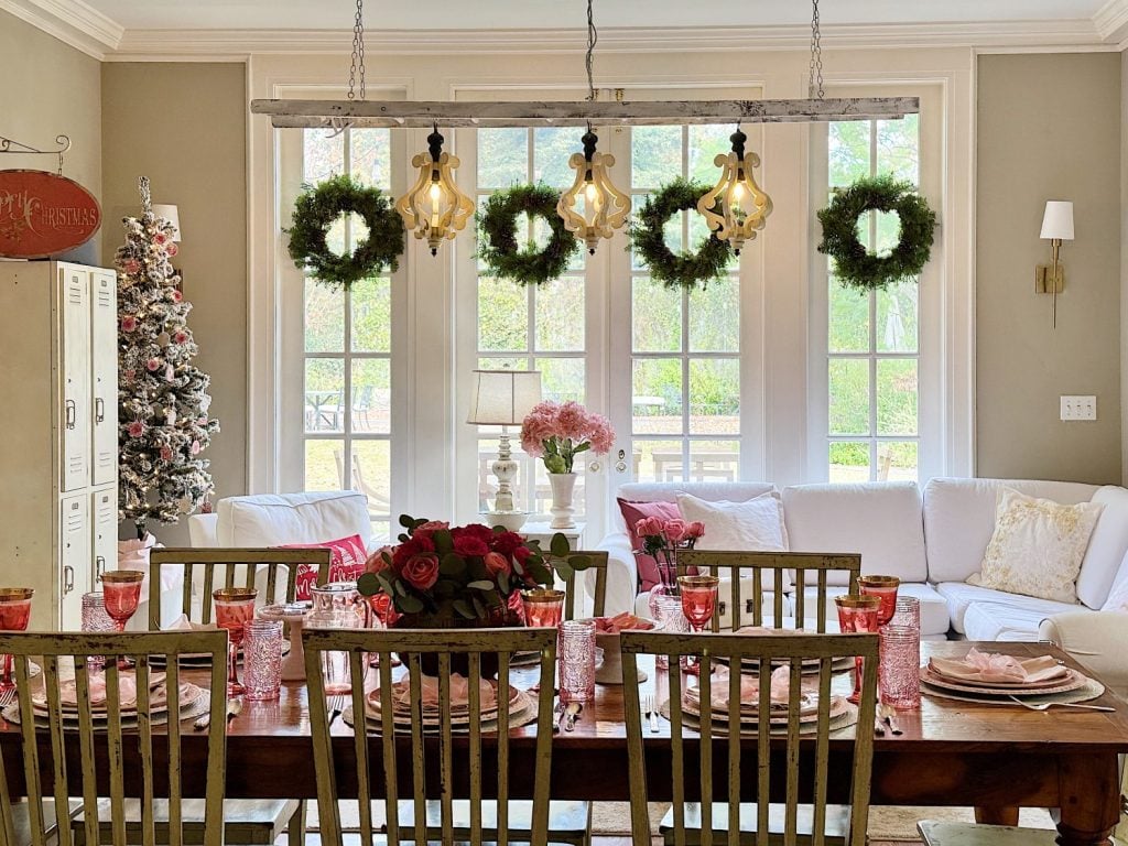 Dining room adorned with floral arrangements and pink glassware. Wreaths hang in front of large windows, and a Christmas tree is visible in the corner.