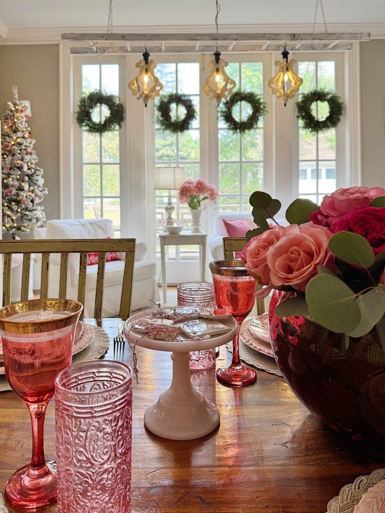 Festively decorated dining room with pink and gold glassware on the table, rose bouquet, and view of a Christmas tree and wreaths through large windows.