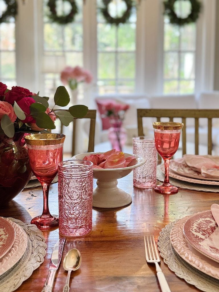 Elegant table set with pink glassware, floral centerpiece, and rose petals, in a bright room with large windows and greenery outside.