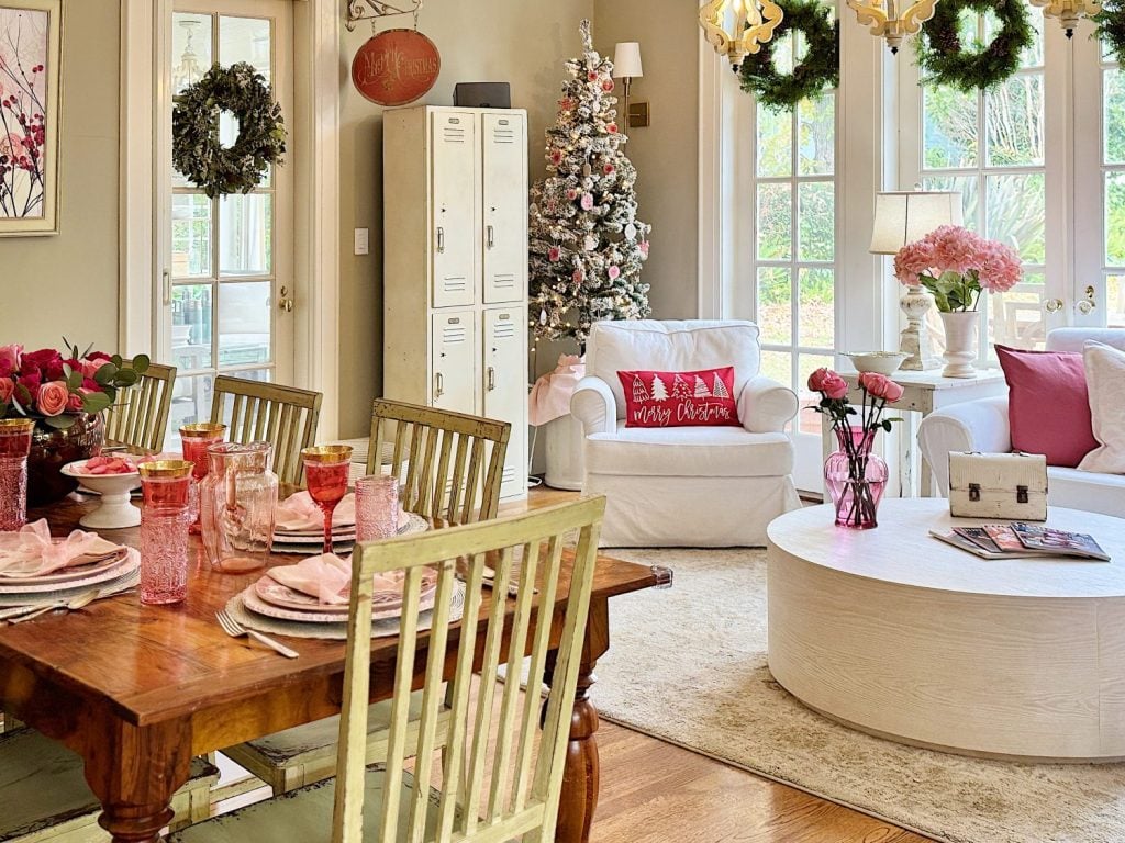 A cozy living and dining space decorated for Christmas, featuring a table set with pink dishware, a tree with ornaments, festive wreaths, and pink and red accents throughout.