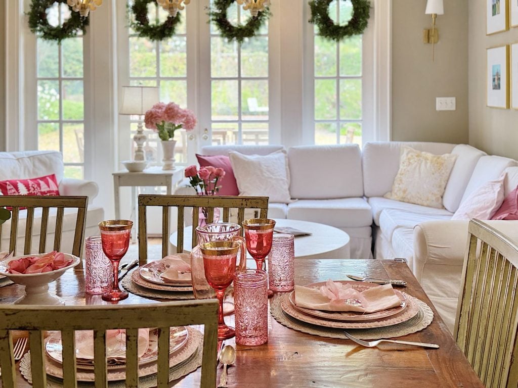 A cozy dining area with pink glasses and plates set on a wooden table. White sofas in the background, decorated with pink pillows and floral wreaths on the windows.