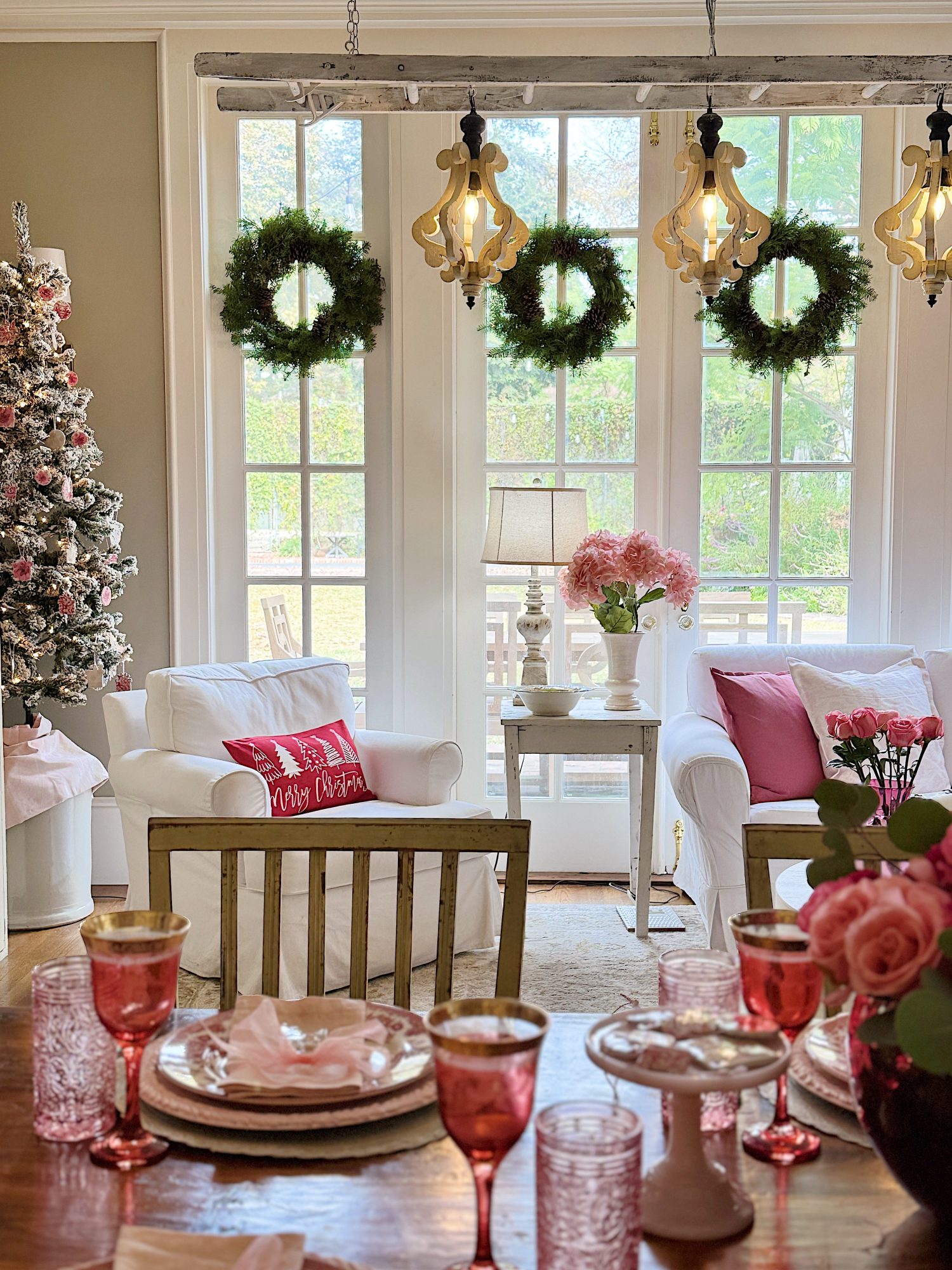 A cozy living room with festive Christmas decor, including a tree, wreaths, and pink accents. A table is set with pink glassware and a white chair has a "Merry Christmas" pillow.