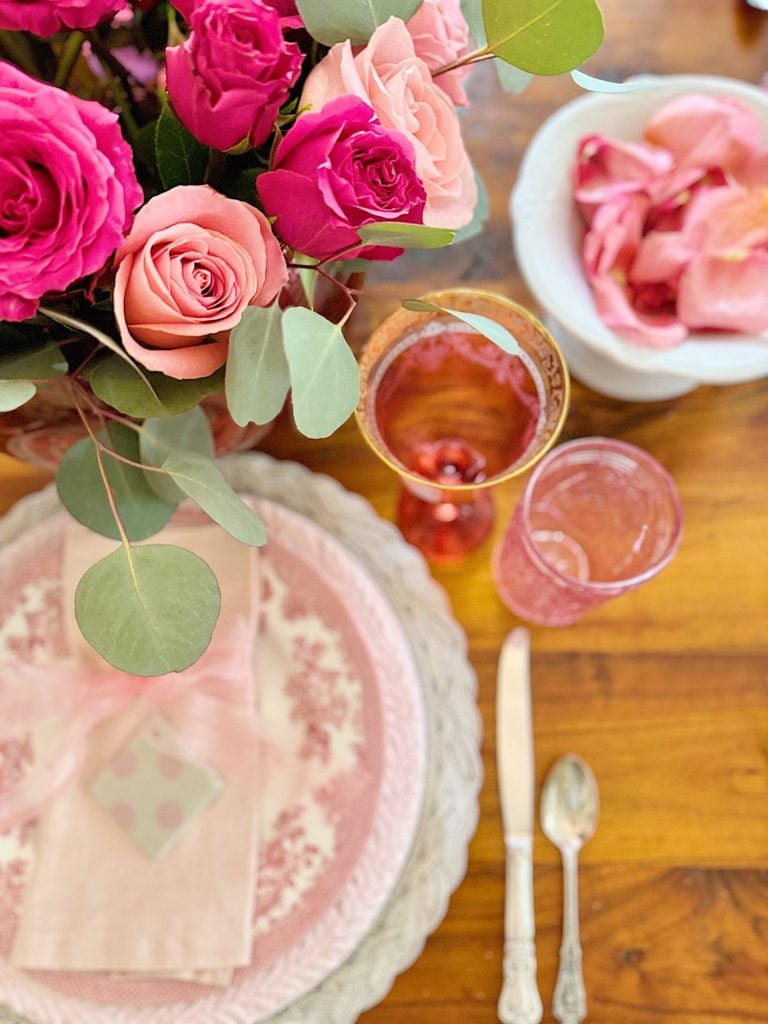 A table setting with pink and red roses, pink glassware, and vintage-style plates. A napkin with a spotted box is placed next to a knife and spoon on a wooden table.