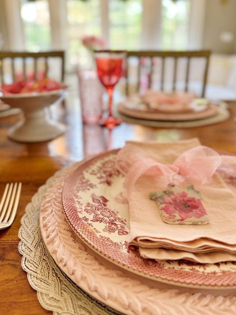 Elegant table setting with pink and floral-patterned plates, pink napkins, a gift tag, and a pink goblet on a wooden table.