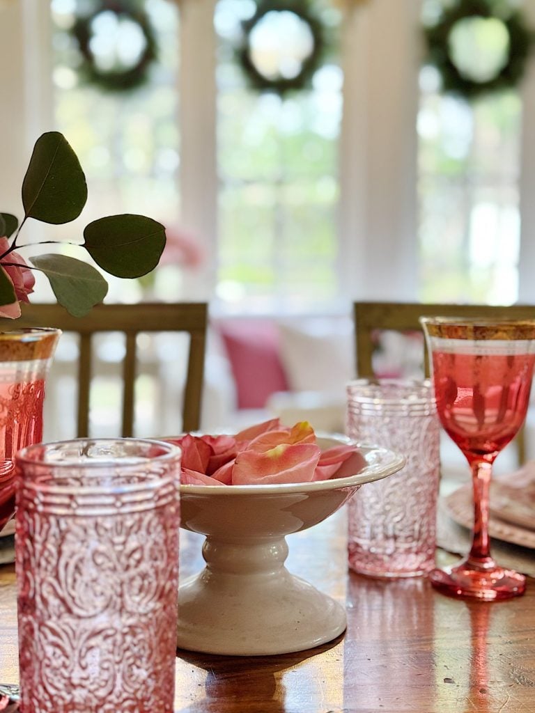 Table setting with pink glasses and a white bowl holding pink petals. Greenery wreaths hang on windows in the background.