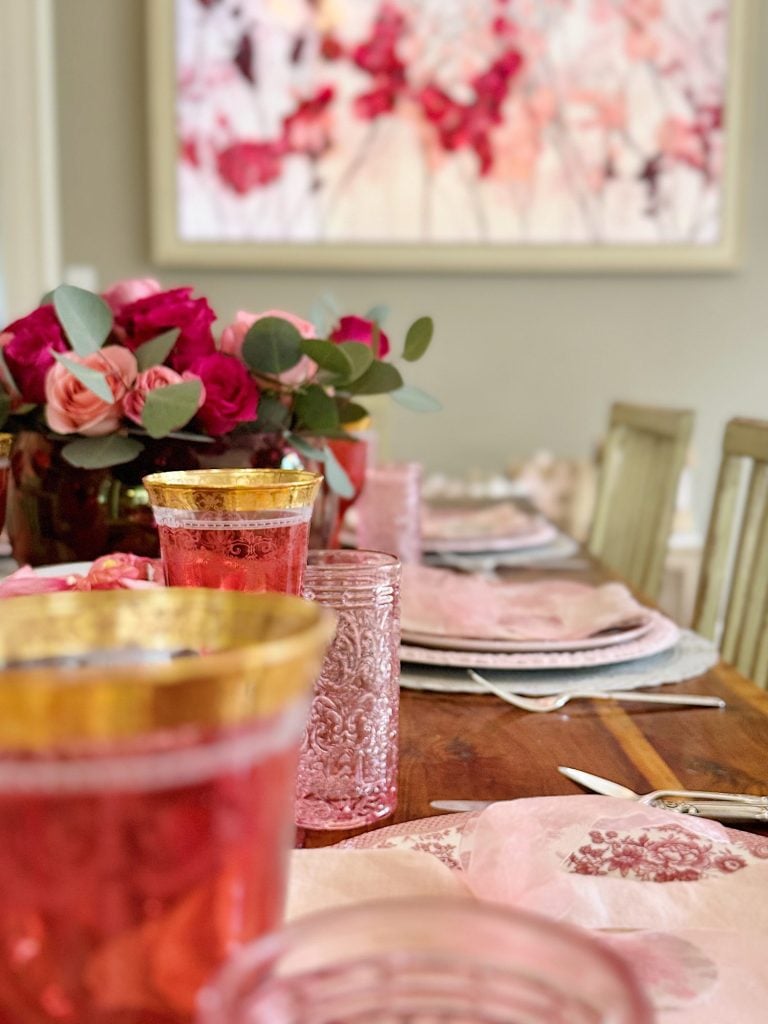 Elegant dining table set with pink and gold glasses, floral arrangements, and pastel plates, beneath a colorful abstract painting.