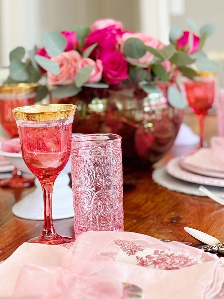 Elegant table setting with pink glassware, gold-rimmed goblet, and floral centerpiece featuring pink and red roses with green leaves on a wooden table.