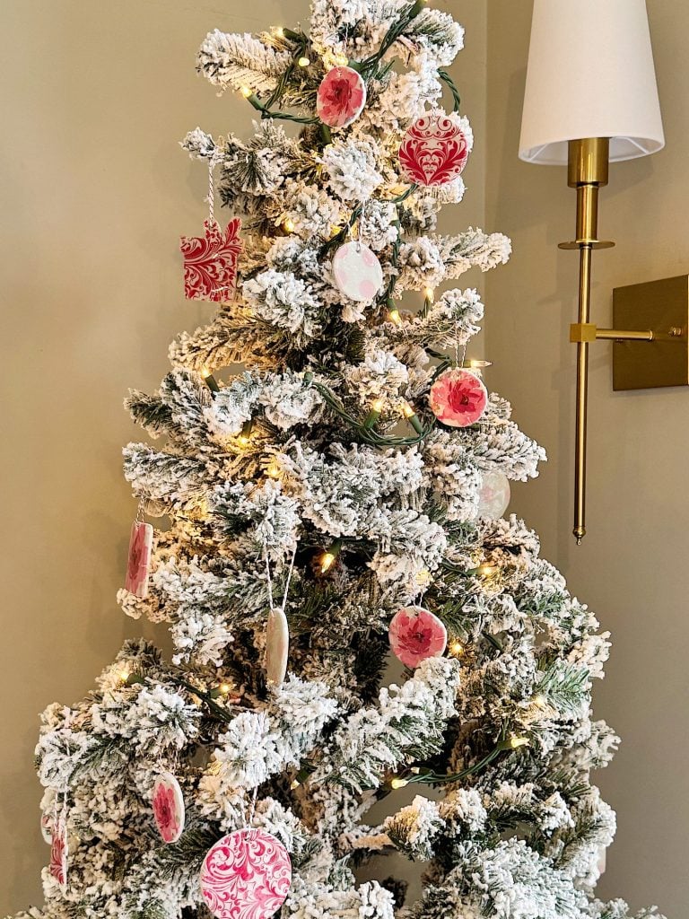 A snow-covered Christmas tree with pink and red ornaments stands next to a brass wall sconce.