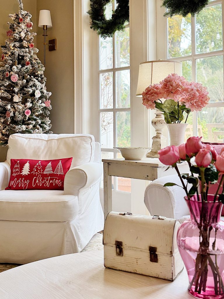 A cozy living room with a decorated Christmas tree, a red pillow on a white chair, pink flowers in a vase, and a window view.