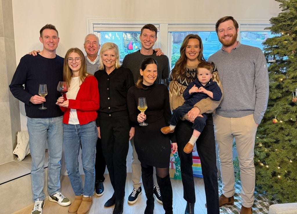 A group of eight adults and one child stand smiling with drinks, next to a decorated Christmas tree indoors.