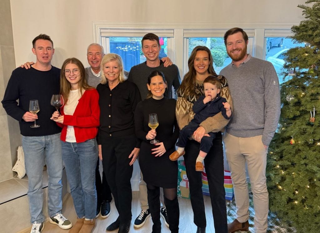 A group of eight people, some holding drinks, stand together indoors near a Christmas tree, smiling at the camera. One woman holds a child in her arms.