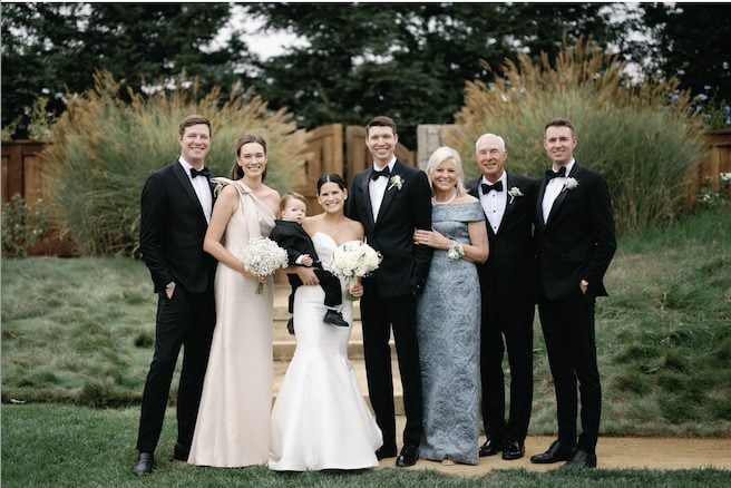 A group of seven people, including a bride holding a child, pose in formal attire outside against a backdrop of greenery and a wooden fence.