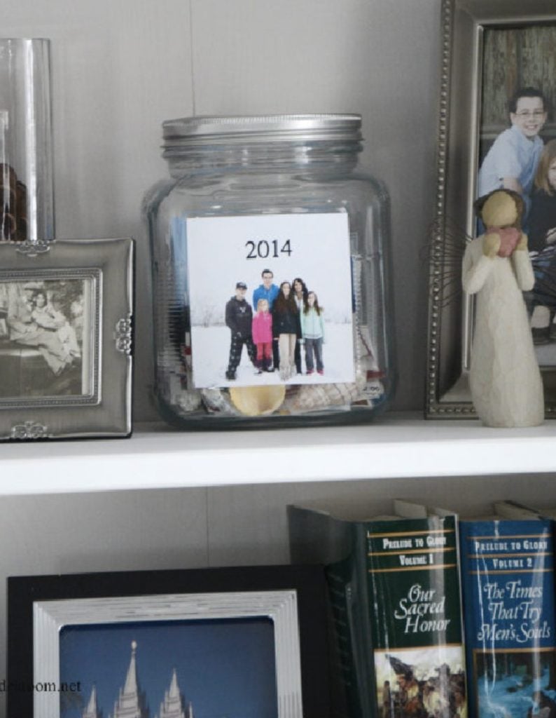 Shelf with framed family photos, a jar labeled "2014" containing a picture of a family, decorative figurine, and books titled "Our Sacred Honor" and "The Times That Try Men's Souls.