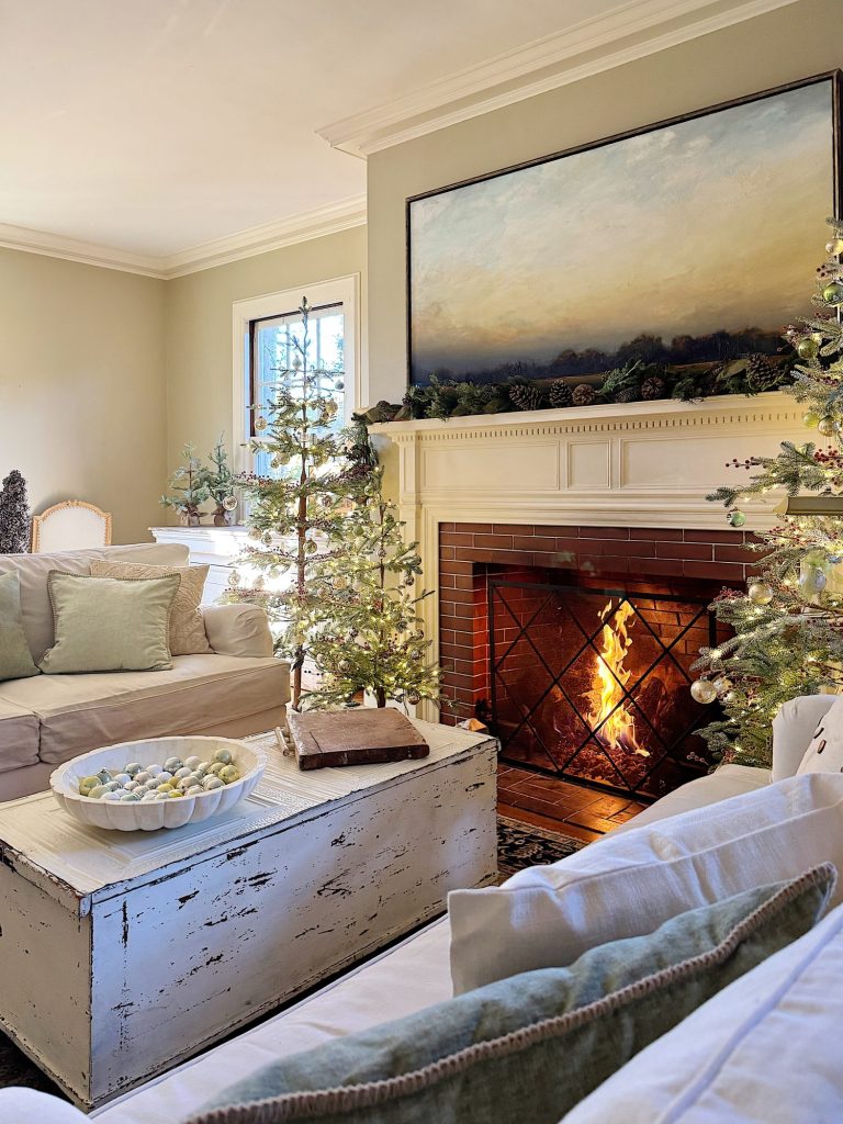 A cozy living room with a lit fireplace, Christmas trees, a white rustic coffee table, and beige sofas adorned with green and white cushions.