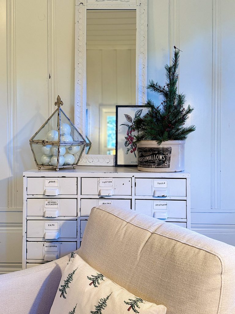 A cozy corner with a beige armchair, a white multi-drawer cabinet, a small potted pine tree, a decorative ornament, and a mirror on the wall.