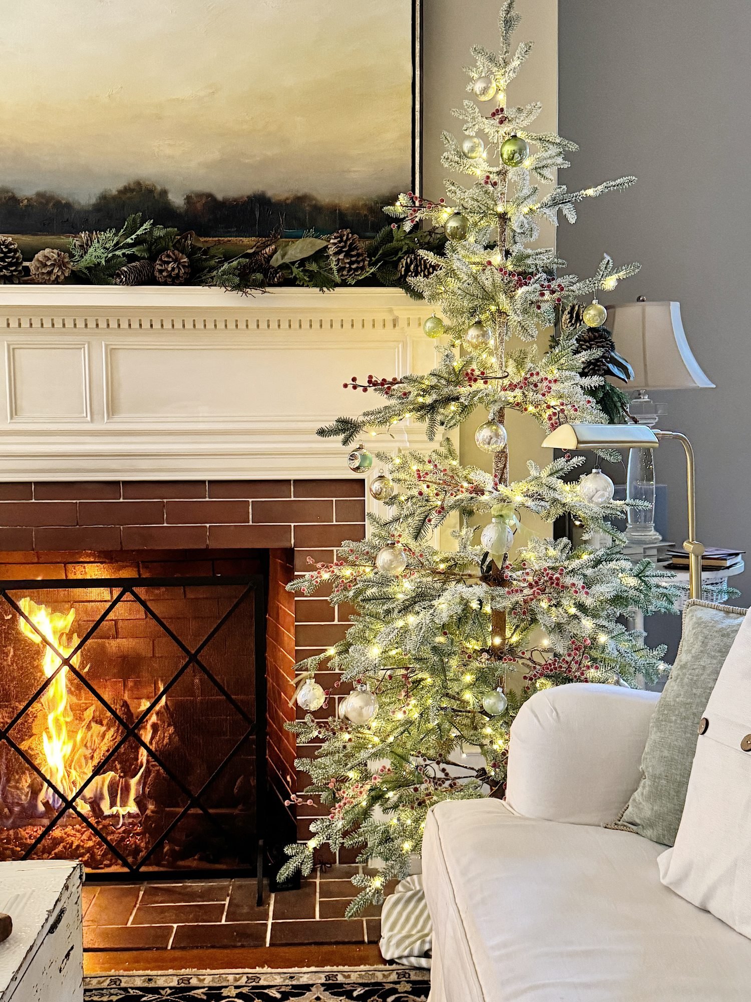 A decorated Christmas tree with lights stands near a burning fireplace in a living room, next to a white sofa. A mantel with pinecone decorations is above the fireplace.