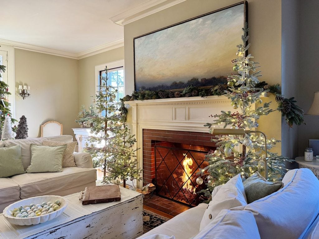 Cozy living room with two lit Christmas trees, a fireplace with a painting above, beige sofas, and a white rustic coffee table with decorations.