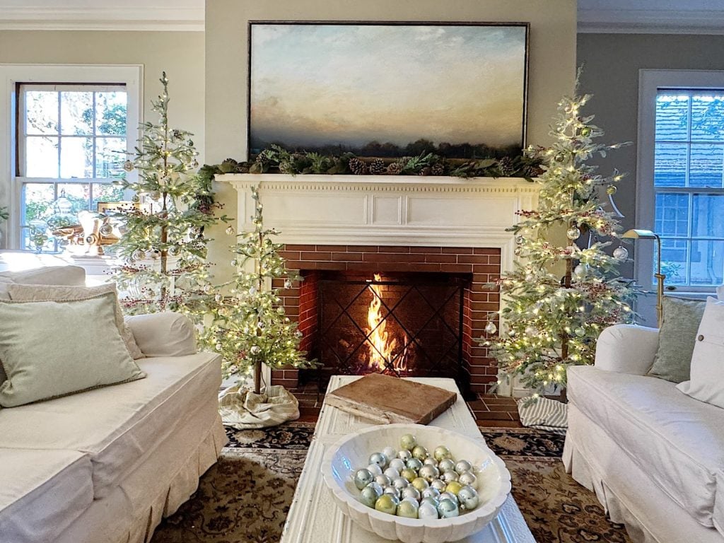 A cozy living room with a lit fireplace, flanked by decorated Christmas trees. A bowl of ornaments sits on a coffee table. A large painting hangs above the mantle.