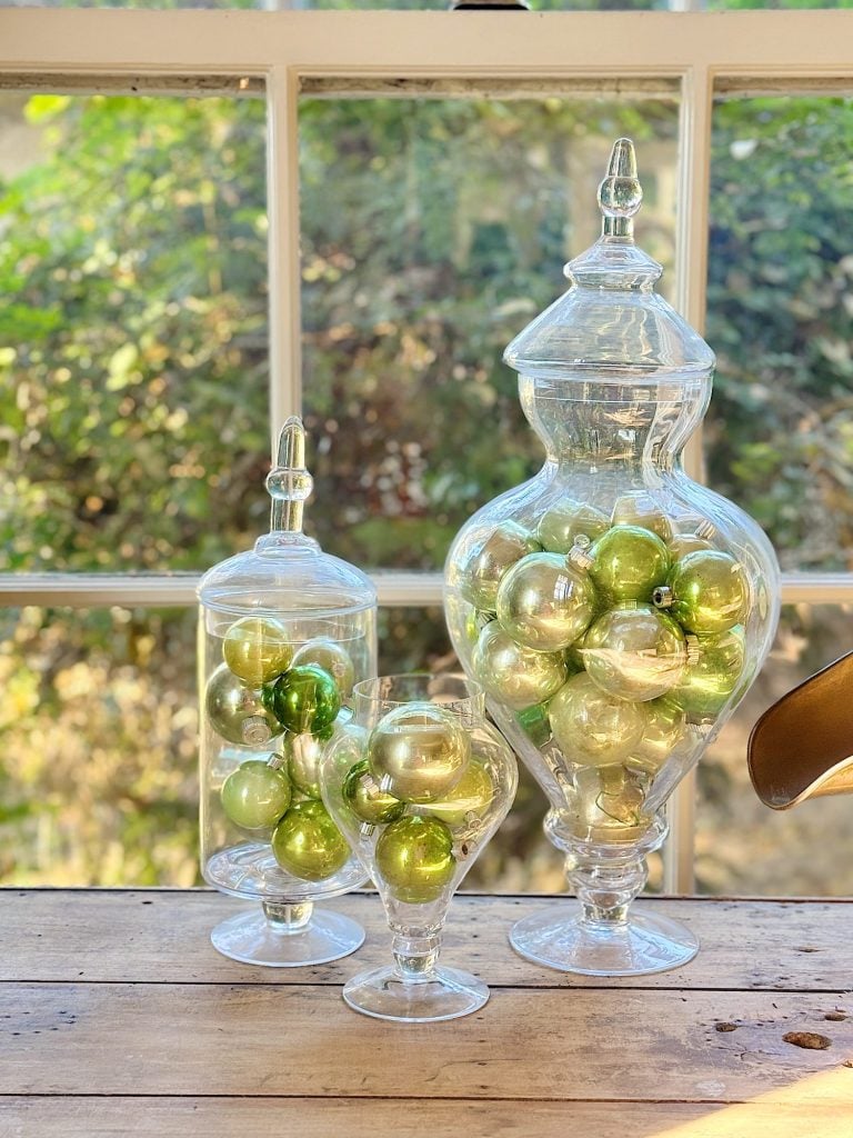 Three glass jars filled with green and silver ornaments are displayed on a wooden table in front of a sunlit window.