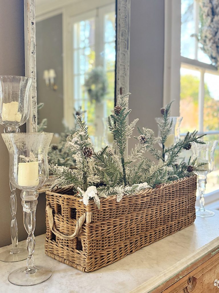 Rattan basket with frosted pine branches and pine cones on a marble surface, flanked by glass candle holders, near a window reflecting outdoor greenery.