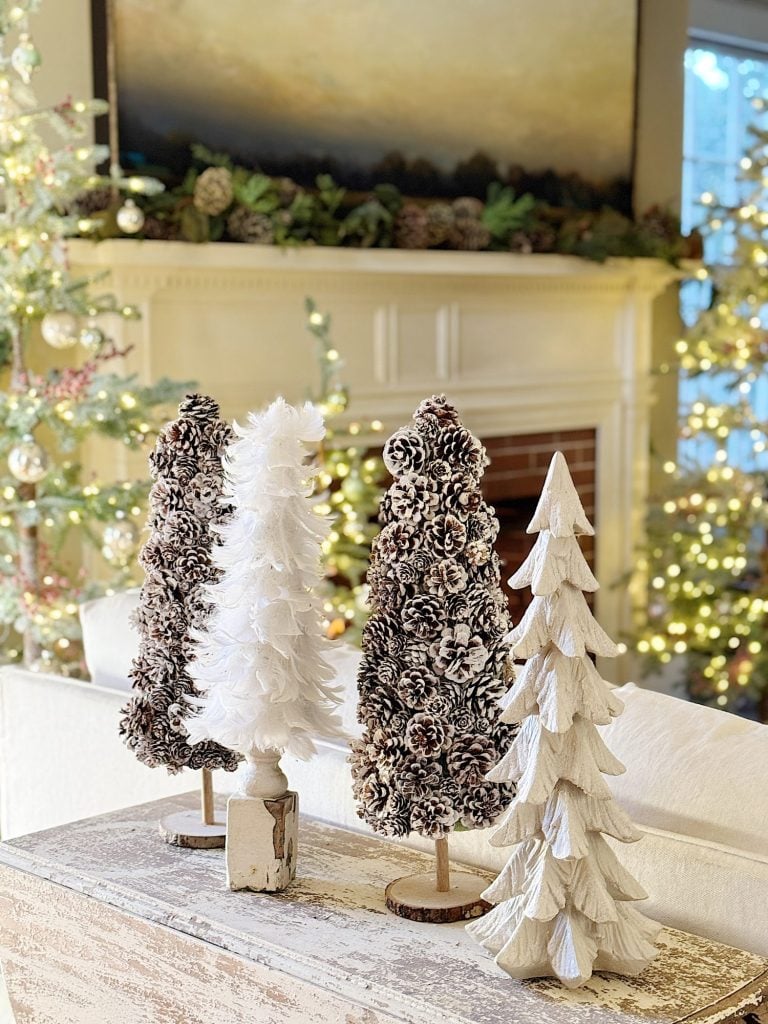 Three decorative trees on a wooden table in front of a fireplace, surrounded by holiday lights and a festive atmosphere.