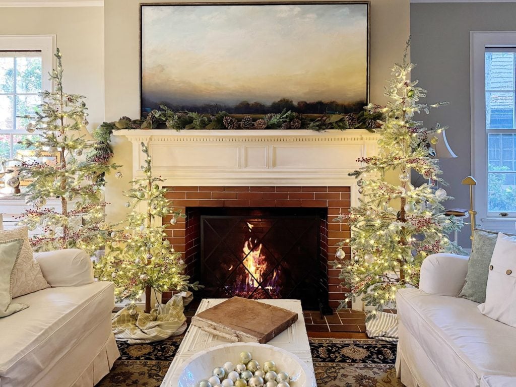 A cozy living room with a lit fireplace, decorated with two small illuminated Christmas trees. A large landscape painting hangs above the mantel. White couches frame the room.