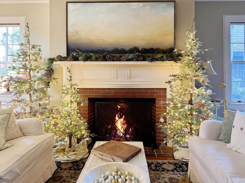 Cozy living room with a lit fireplace, adorned with holiday decorations. Two slim Christmas trees with lights flank the mantel, which is topped with a large painting and pinecone garland.