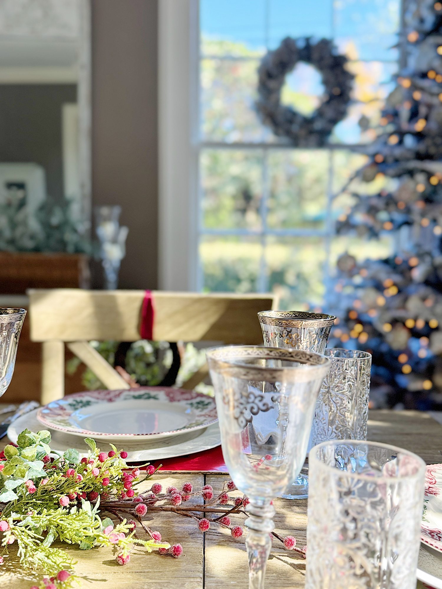 Festive table setting with glassware, a decorative plate, and a sprig of berries. A Christmas tree and wreath are visible in the background near a window.