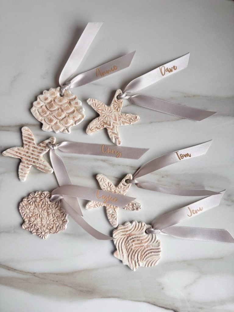 Six textured white clay ornaments with ribbon loops, each labeled in gold script, arranged on a white marble surface—perfect for personalized table setting ideas.