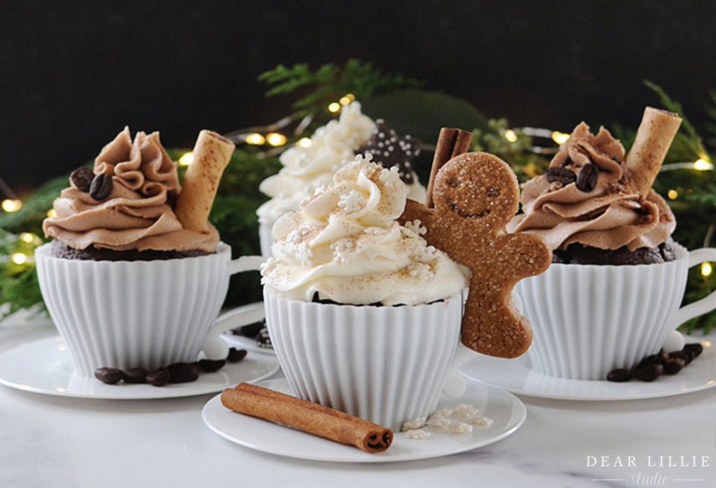 Four decorated cupcakes in white cups, topped with frosting, waffle rolls, coffee beans, and a gingerbread cookie. Cinnamon sticks are placed in front. Holiday lights in the background.