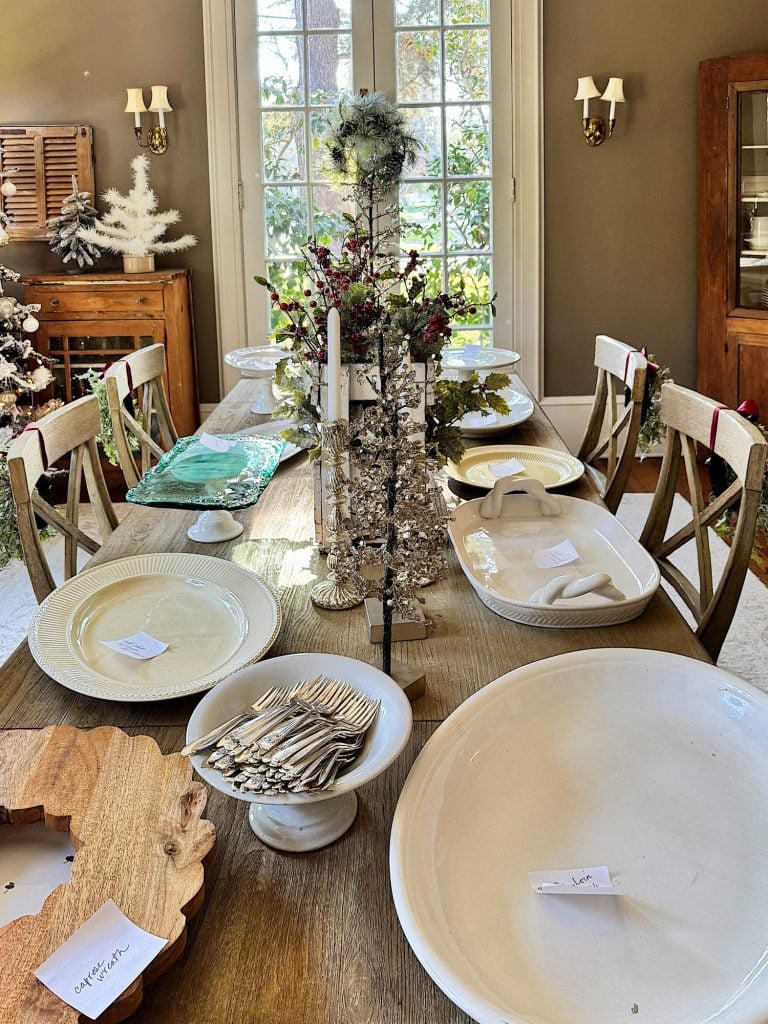 A dining table set with plates, serving dishes, utensils, and a decorative centerpiece. The room is warmly lit and features large windows and wooden chairs.