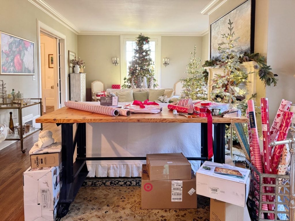 Room with a large table covered in gift wrapping materials and boxes. Christmas trees and decorations are in the background.