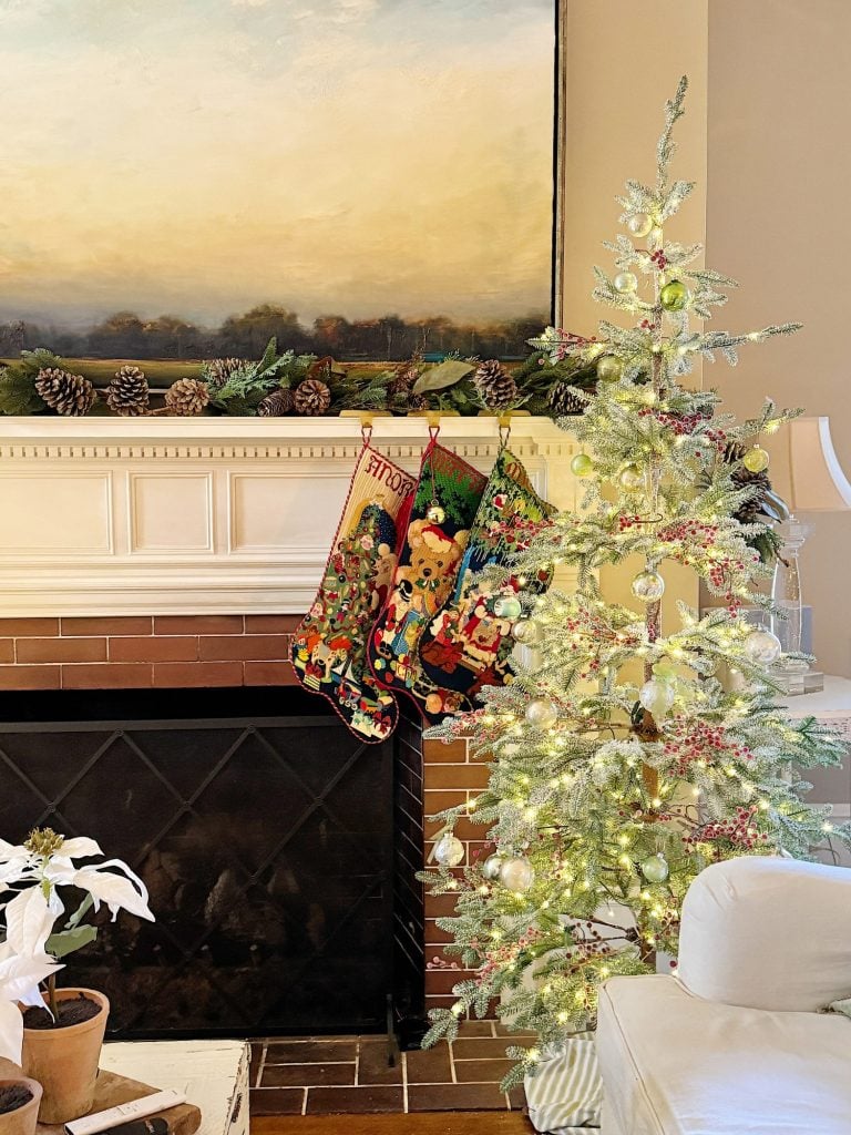 A decorated Christmas tree sits by a fireplace with three festive stockings hanging. The mantle is adorned with pinecones, and a large landscape painting hangs above.