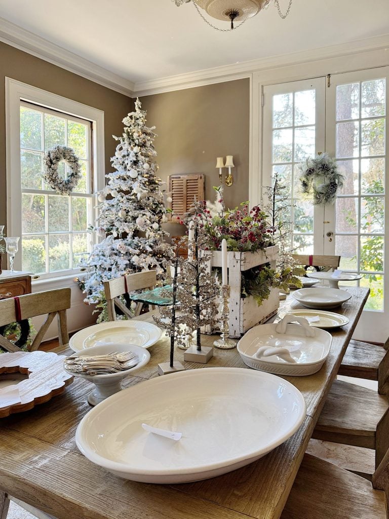 Decorated dining room with a wooden table set for a meal, featuring white plates, a centerpiece, small decorative trees, and a flocked Christmas tree by a window.