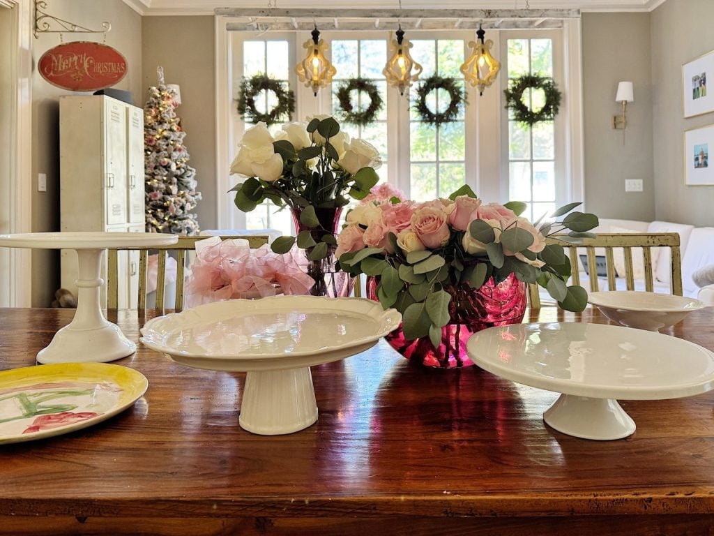 Dining table display with cake stands, floral arrangements in vases, and a "Merry Christmas" sign. Christmas tree and wreaths visible in the background near tall windows.