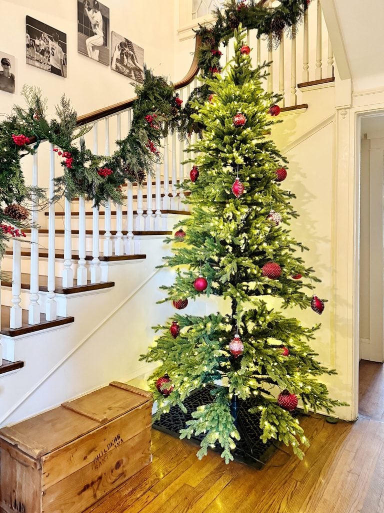 A Christmas tree with red ornaments stands by a staircase decorated with garland in a warmly lit hallway.