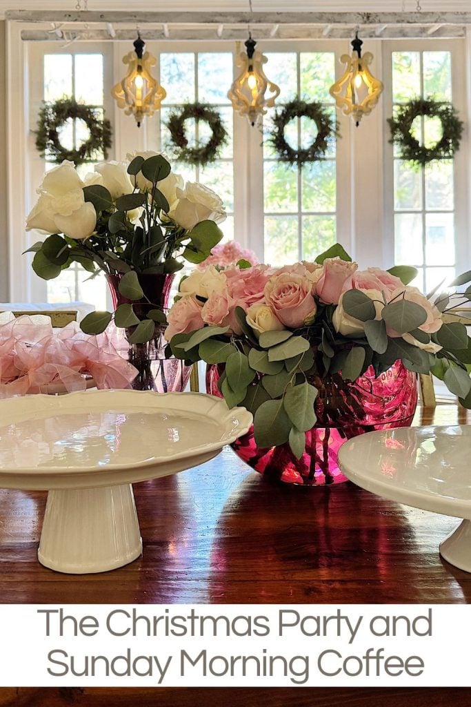 Floral arrangements on a wooden table with cake stands, under hanging lights. Wreaths decorate the windows in the background.