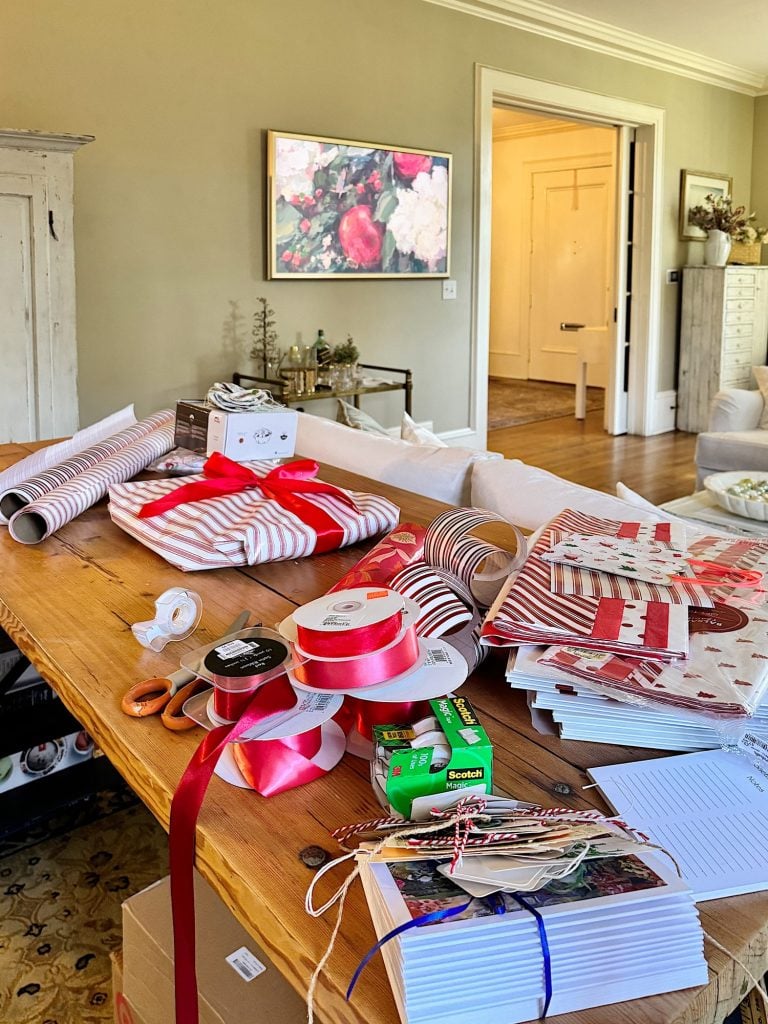 A table cluttered with assorted wrapping papers, ribbons, scissors, tape, and neatly stacked envelopes in a living room with artwork on the wall in the background.