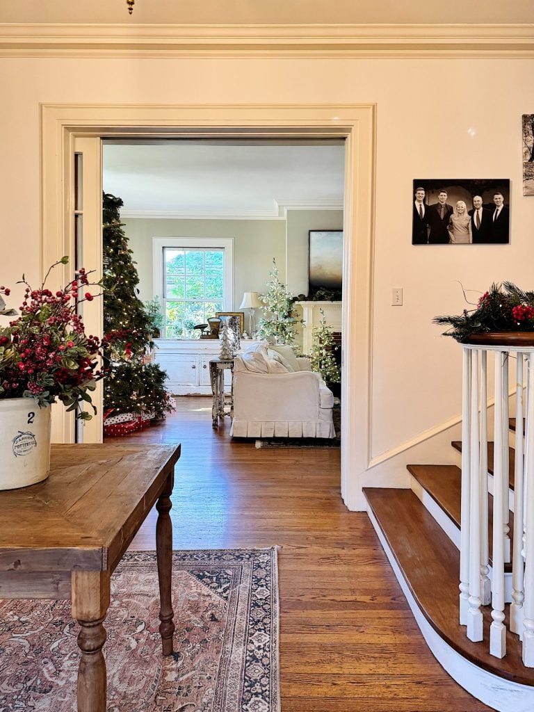 A cozy living room decorated for Christmas, featuring a large tree, floral arrangements, and a view of the outdoors through a window. There's a wooden table and staircase in the foreground.