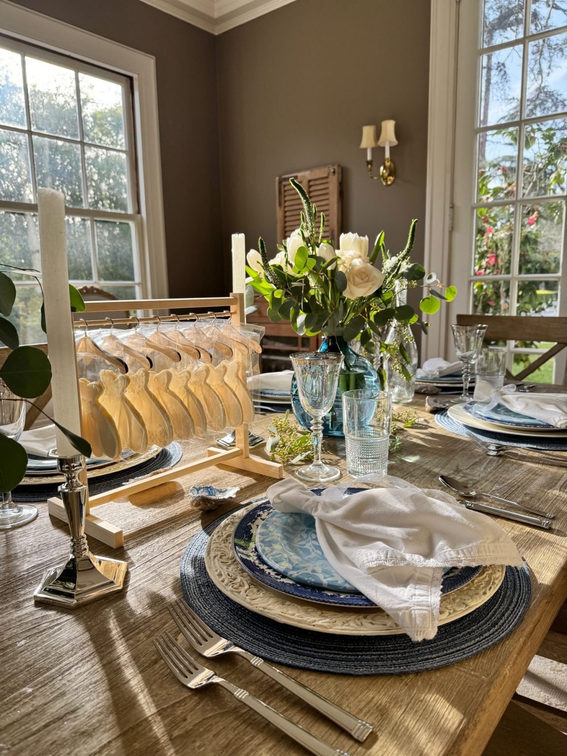 Table set with blue and white plates, wine glasses, white napkins, a vase of flowers, candles, and a wooden rack with bagels, in a sunlit dining room.