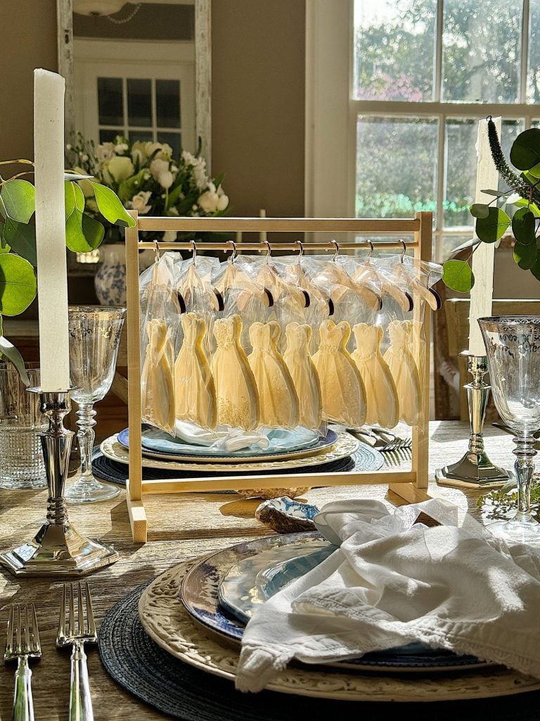 Elegant dining table setup with candles, glassware, and decorative plates. A centerpiece displays cookie favors shaped like dresses, hanging on a wooden rack.