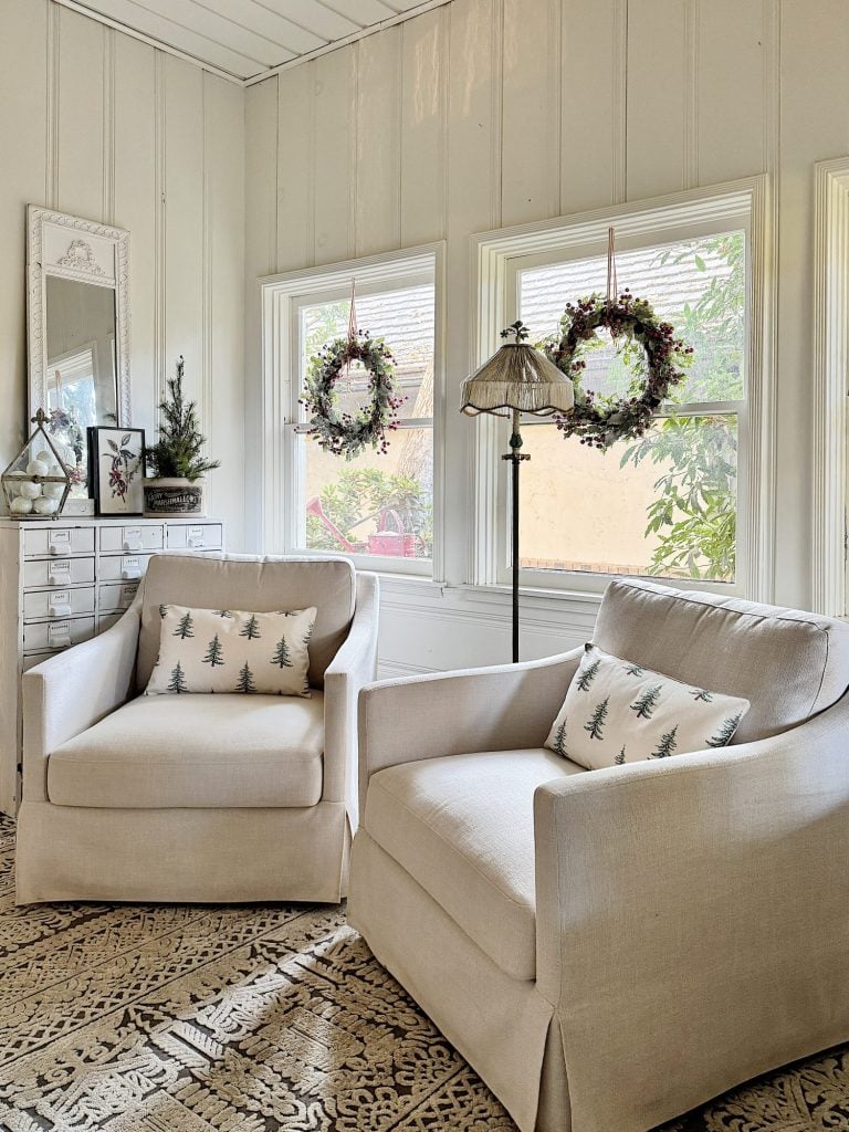 Two beige armchairs with tree-patterned pillows in a bright room with three windows. Windows are decorated with floral wreaths. A floor lamp and a chest of drawers are in the background.