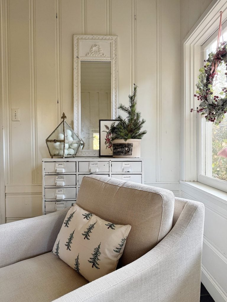 A cozy room with a beige chair, a pillow with tree designs, a small cabinet, a mirror, and holiday decorations including a wreath and potted plant. Large window with outdoor view.