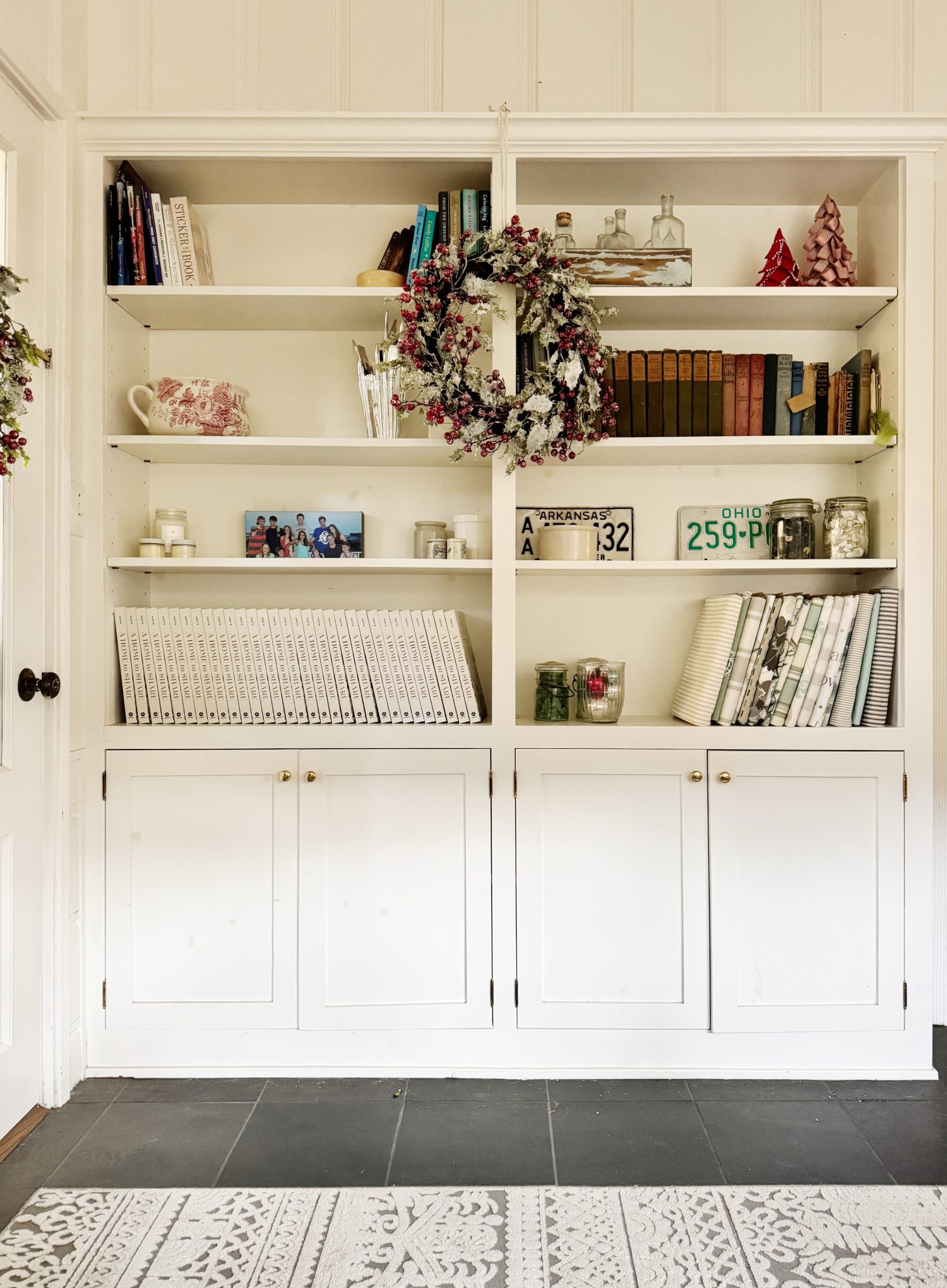 White built-in bookshelves with books, a family photo, decorative wreaths, small ornaments, and vintage decor. Lower cabinets have closed storage. Gray floor and patterned rug visible.